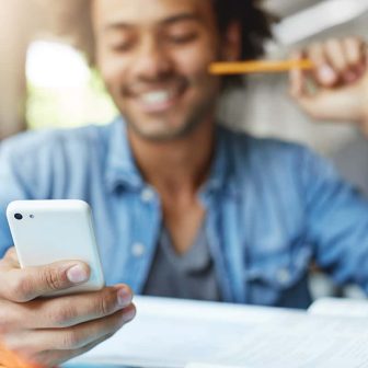 People, lifestyle, technology and communication concept. Handsome bearded dark-skinned male student wearing blue shirt using cell phone, browsing newsfeed via social networks, laughing at memes